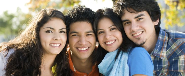 Four latino students -two girls and two boys- smiling directly at the camera, with a leafy background